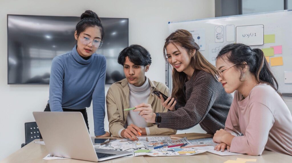 Four young professionals working together around a table with a laptop and design sketches, discussing ideas in a modern office with a whiteboard in the background.