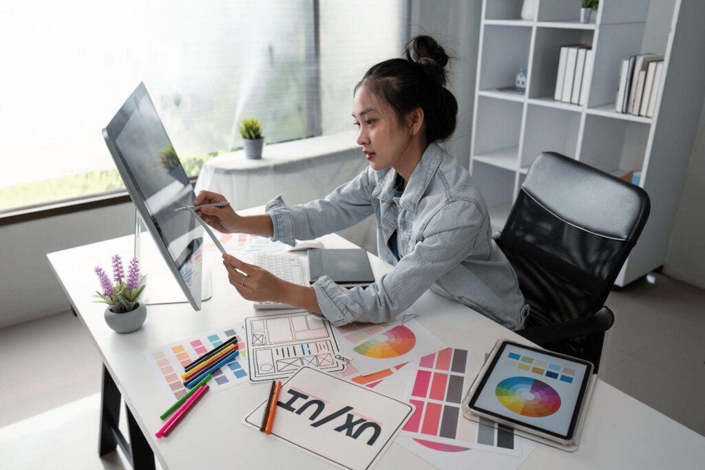 Young designer working at a desk with a computer, reviewing design sketches and color palettes in a bright, modern office.