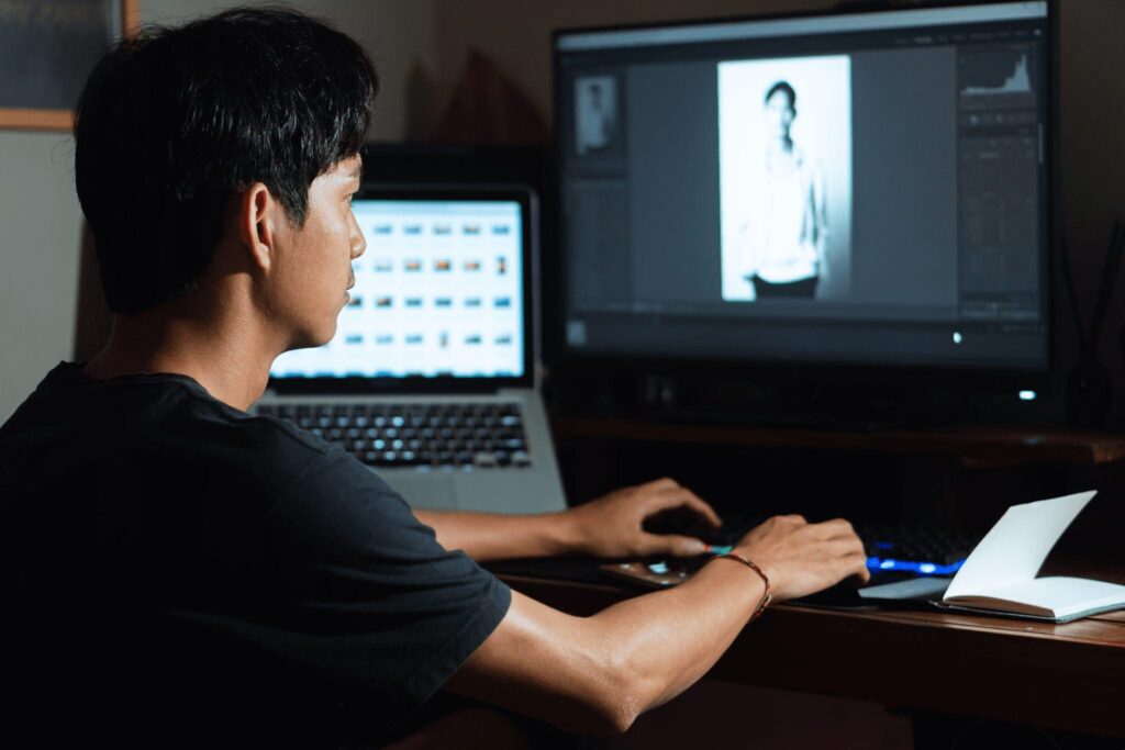 Man working on photo editing software at a desk, focused on adjusting an image on a computer screen in a dimly lit room.