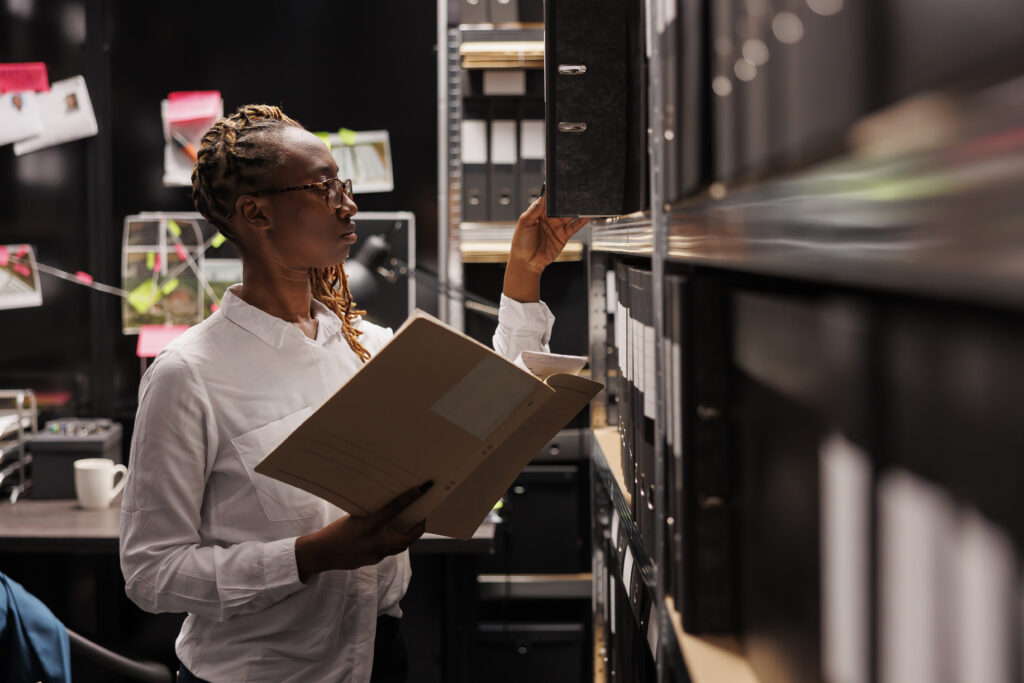 Police investigator studying archival crime case materials, working overtime in detective office room. African american policewoman searching evidence records and taking file from shelf